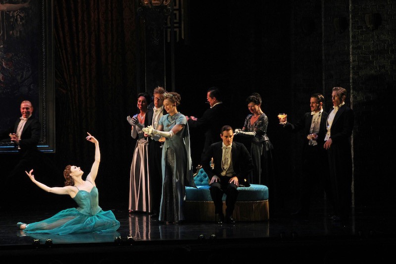 The company in evening wear surrounds Sara Mearns as Victoria Page who is sitting ion the grounds with her arms outstretched. She wears an emerald dress.